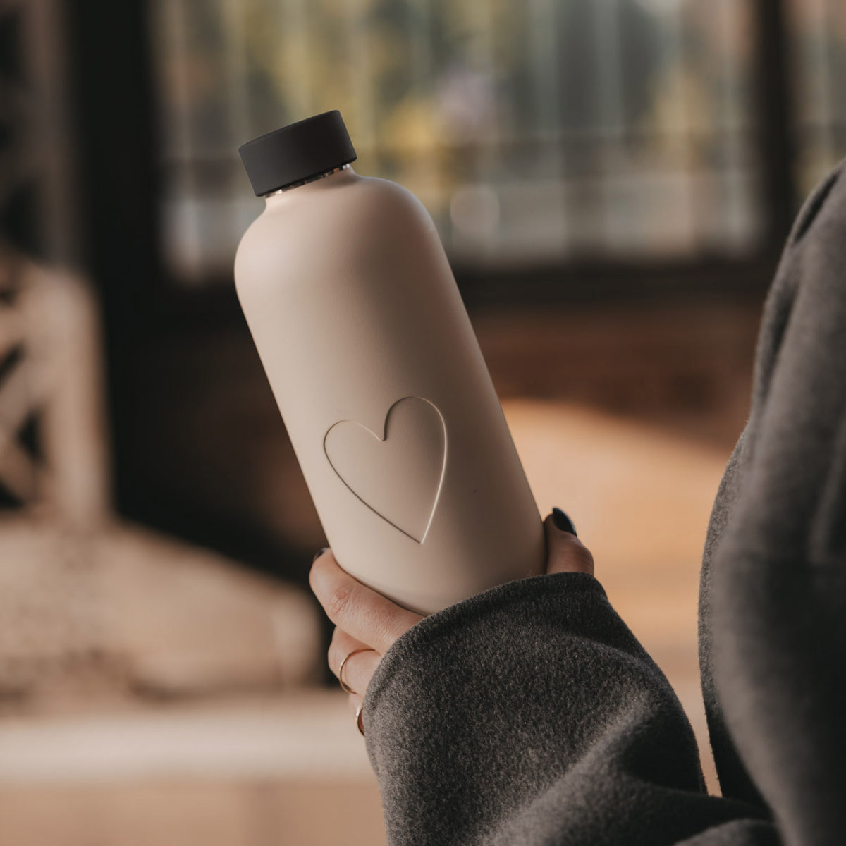 Nahaufnahme einer cremefarbenen Trinkflasche mit Herzmotiv, die eine Frau in der Hand hält.