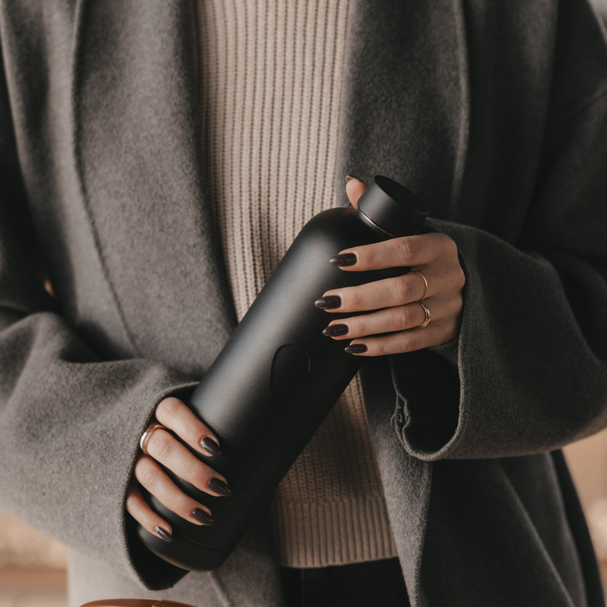 Frau im grauen Mantel hält eine schwarze 1000 ml Trinkflasche mit Herzmotiv und Schraubverschluss in beiden Händen.