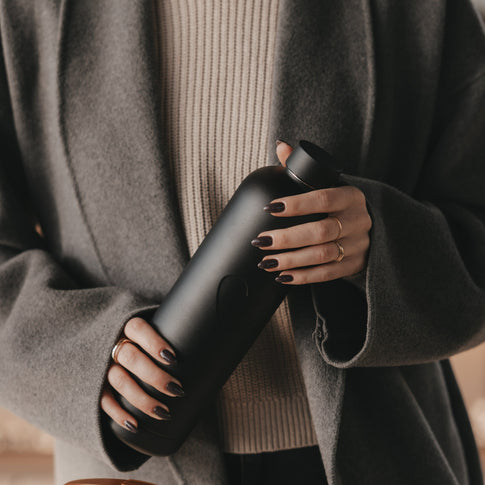 Frau im grauen Mantel hält eine schwarze 1000 ml Trinkflasche mit Herzmotiv und Schraubverschluss in beiden Händen.