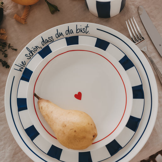 Gestapeltes Geschirr: Platzteller „Wie schön, dass du da bist“ in Dunkelblau, darauf ein blau gestreifter, welliger Speiseteller und ein Frühstücksteller mit rotem Herz, umgeben von Tasse und Kräutern auf hellem Leinentisch.