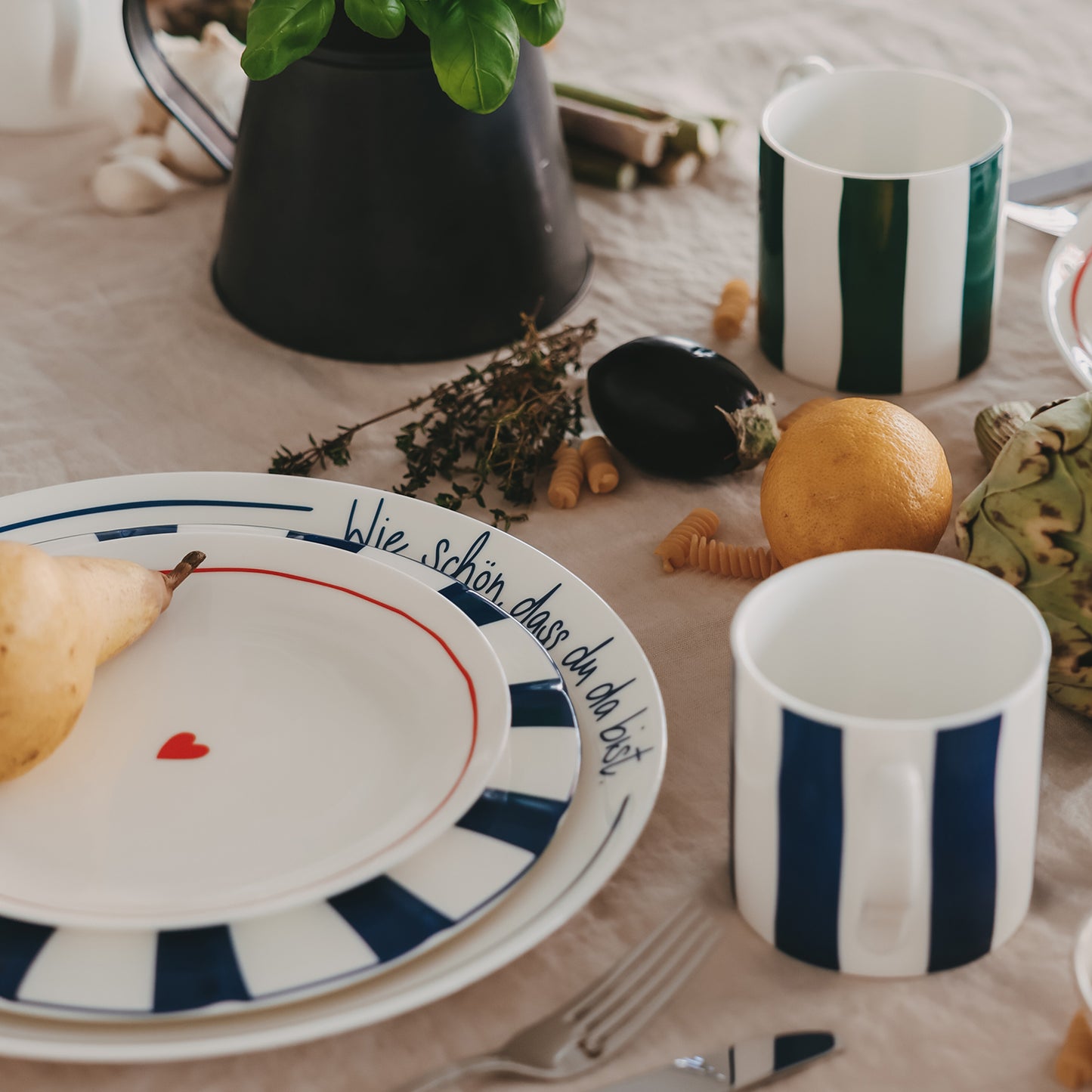 Detailansicht eines gedeckten Sets: Platzteller mit Schriftzug „Wie schön, dass du da bist“, darauf ein blau gestreifter Speiseteller und ein Frühstücksteller mit rotem Herz, dekoriert mit Birne auf hellem Tisch mit Essensdekoration.