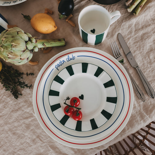 Gestapelte Porzellanteller – kleiner Teller mit grünem Streifenrand, Speiseteller „Pasta Club“ in blau und Platzteller mit rotem Rand, kombiniert mit Tasse und frischen Zutaten auf Leinentischdecke.