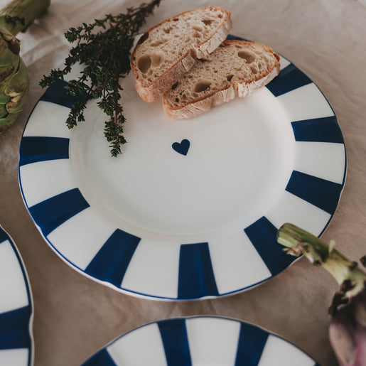 Porzellan Speiseteller mit welligem Rand, blauem Streifenmuster und kleinem Herz, auf Leinentischdecke mit Brot und Kräutern arrangiert