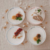 4 „pasta club“ Speiseteller in blau, hellblau, grün und rot, angerichtet mit Pasta, Brot und Mozzarella, umgeben von Knoblauch und trockenen Nudeln auf Leinentisch.