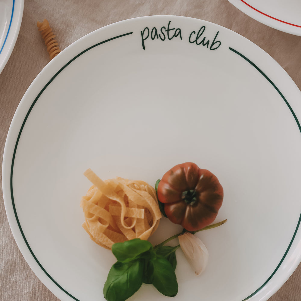 Speiseteller „pasta club“ mit grünem Rand, angerichtet mit Pasta-Nest, dunkler Tomate, Basilikum und Knoblauch auf hellem Leinentisch.