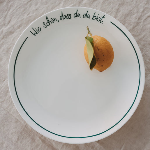 Weißer Platzteller mit grüner Schrift „Wie schön, dass du da bist“ mit Zitrone, auf hellem Leinentisch.
