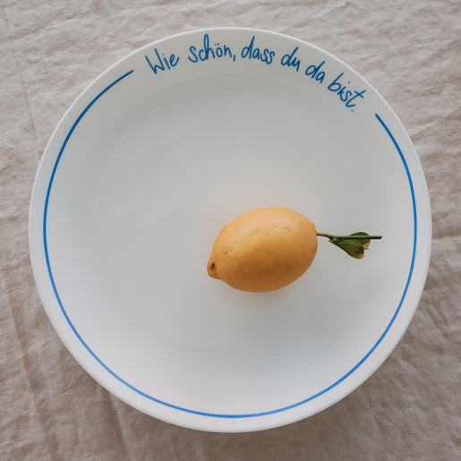 Weißer Platzteller mit hellblauer Schrift „Wie schön, dass du da bist“ mit Zitrone, auf hellem Leinentisch.