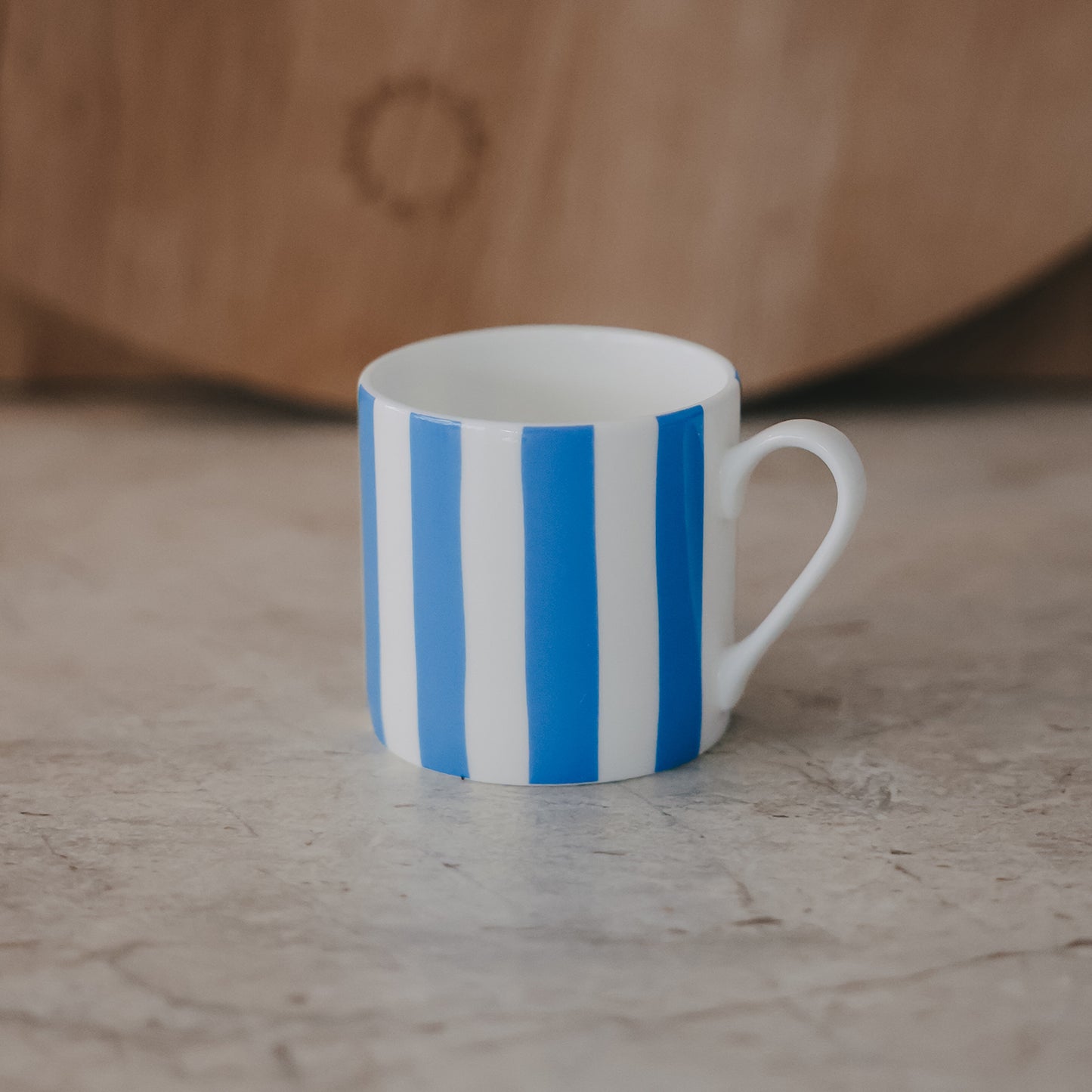 Kleine Tasse mit hellblauen, vertikalen Streifen und zartem Henkel vor neutralem Hintergrund mit Holzbrett.