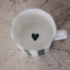 Blick in die Tasse mit weißem Inneren und kleinem grünem Herz am Boden, auf heller Steinoberfläche.