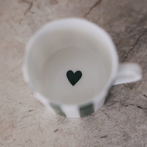 Blick in die Tasse mit weißem Inneren und kleinem grünem Herz am Boden, auf heller Steinoberfläche.