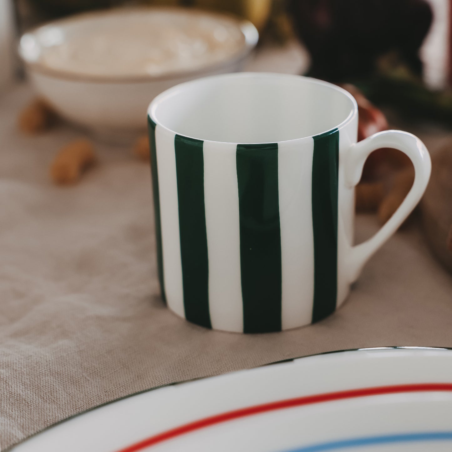 Kleine Porzellan-Tasse mit grünen Streifen auf gedecktem Tisch, im Vordergrund Teller mit farbigem Rand.