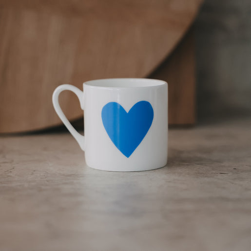 Kleine weiße Tasse mit großem hellblauen Herz frontal auf heller Steinfläche, weicher Hintergrund.