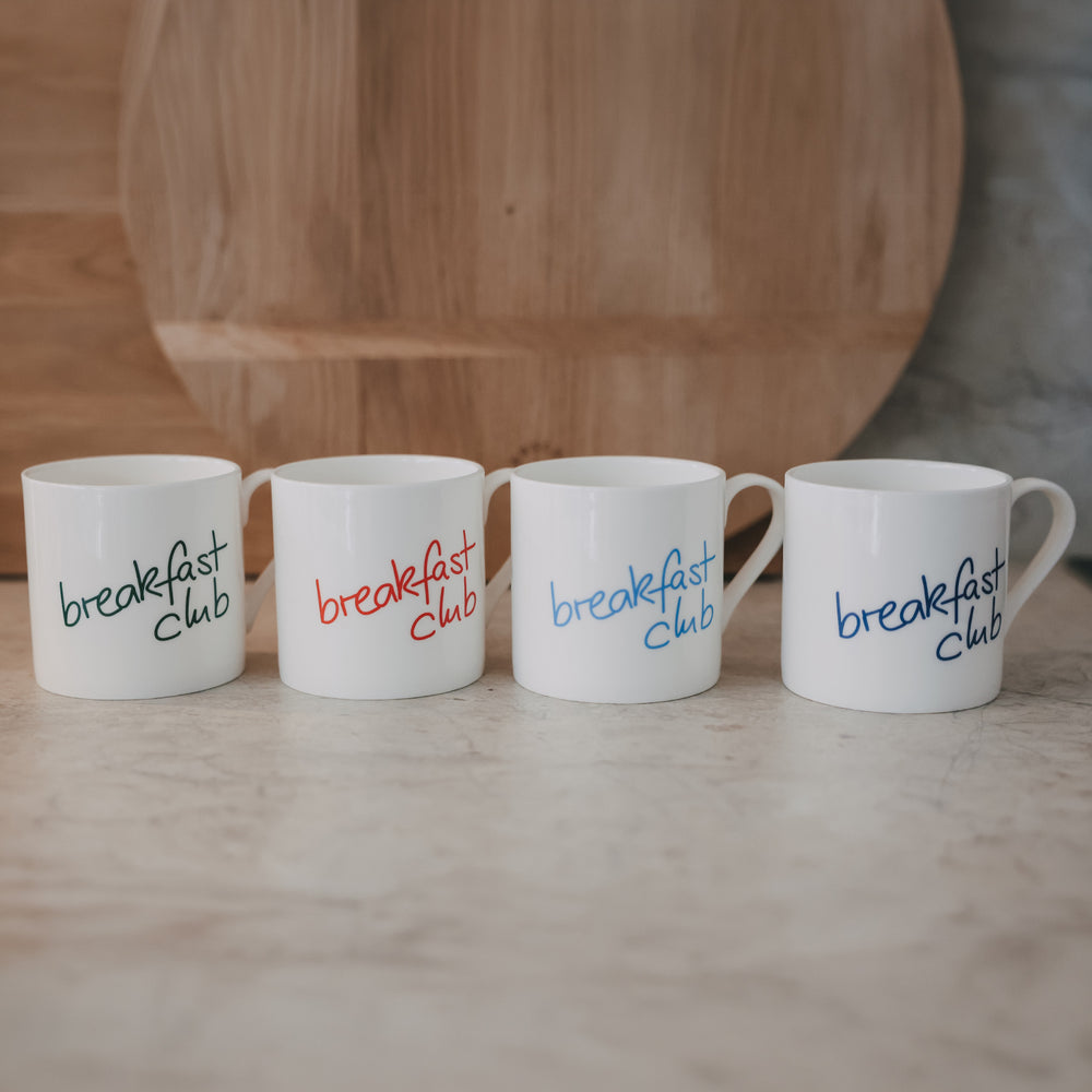 Vier weiße Tassen mit „breakfast club“-Schrift in Grün, Rot, Hellblau und Blau nebeneinander auf hellem Tisch vor Holzbrett