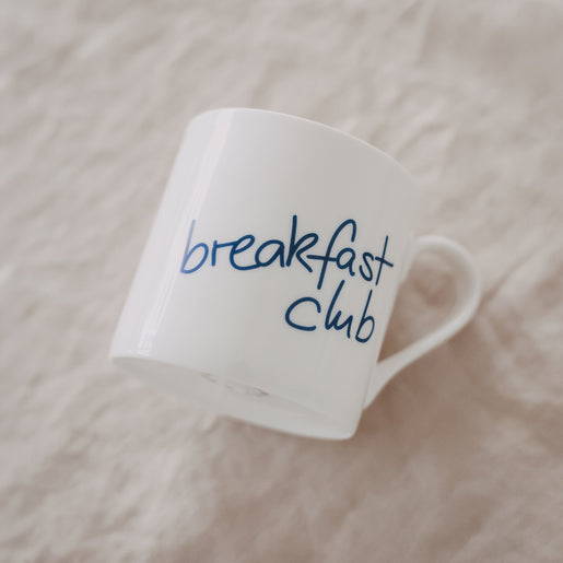 Schräg liegende weiße Tasse mit blauem „breakfast club“-Schriftzug auf hellem Untergrund