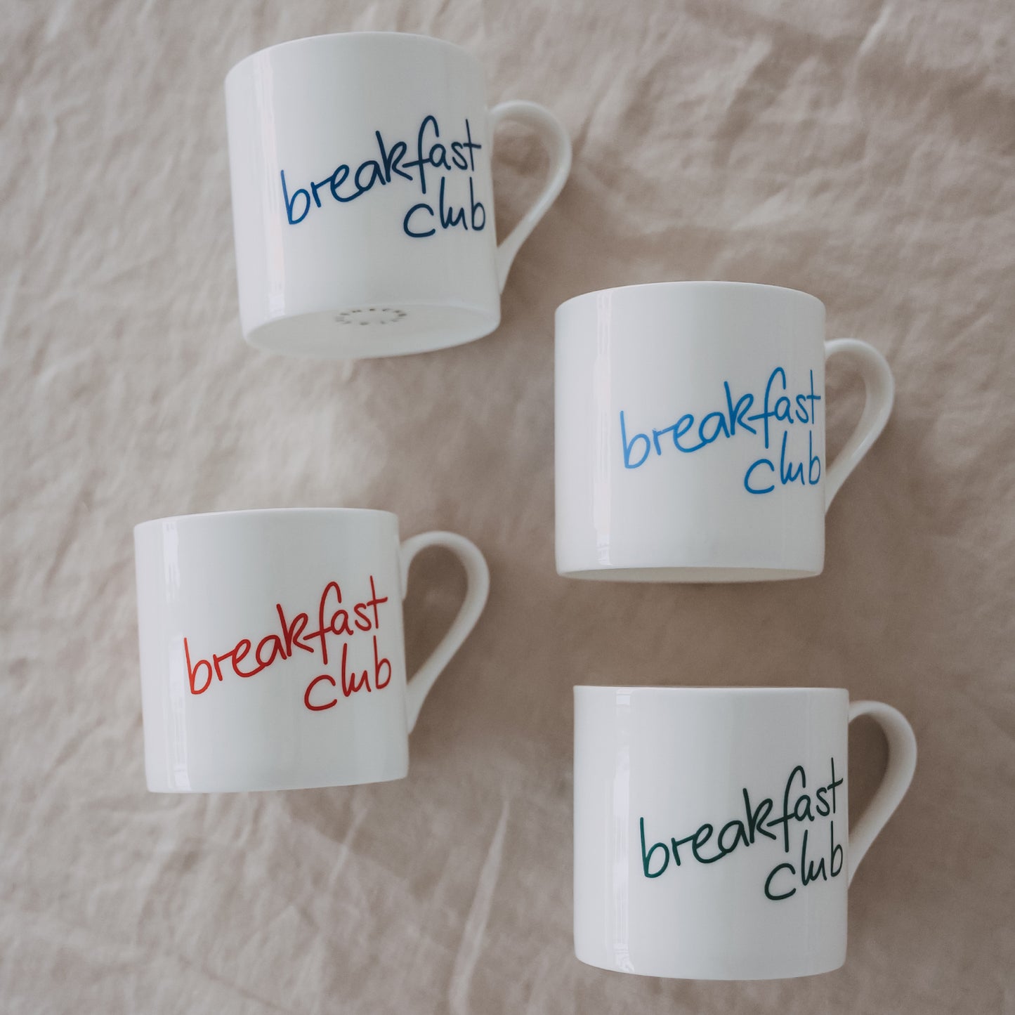 Mehrere weiße „breakfast club“-Tassen in verschiedenen Farben auf hellem Stoff arrangiert, Detailansicht