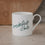 Weiße Tasse mit grünem „breakfast club“-Schriftzug vor warmem Hintergrund