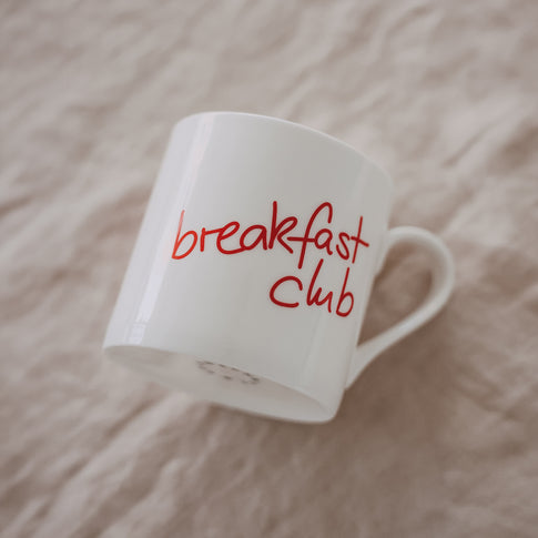 Schräg liegende weiße Tasse mit rotem Schriftzug „breakfast club“ auf hellem Untergrund