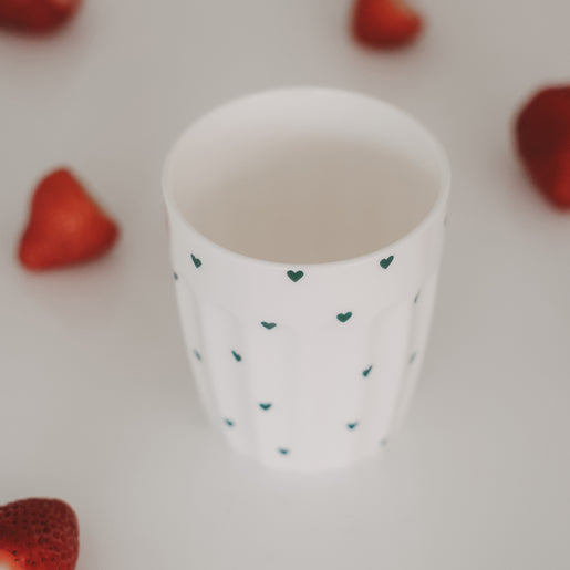 Keramikbecher mit grünen Herzen, umgeben von frischen Erdbeeren auf hellem Hintergrund