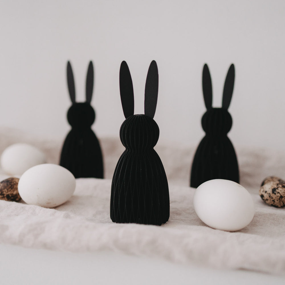 Drei schwarze Papierhasen als Osterdeko, stehend zwischen weißen und gesprenkelten Eiern auf hellem Stoff.