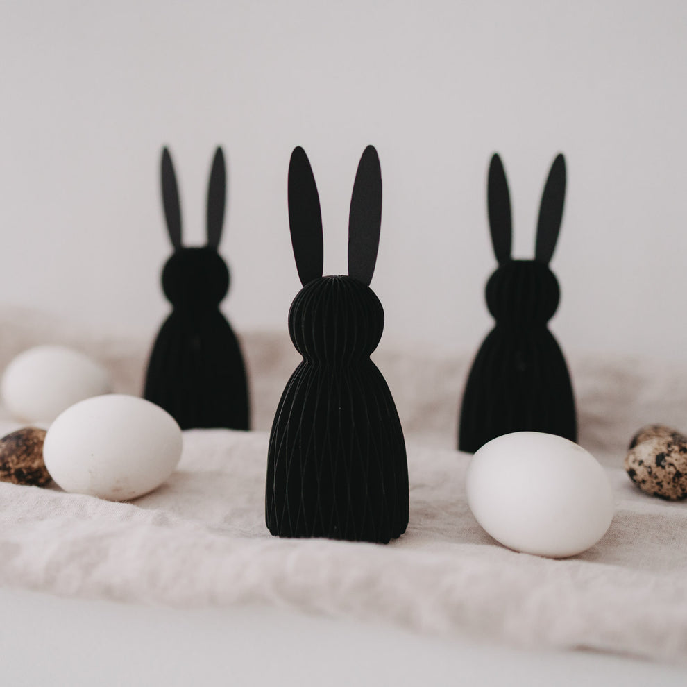 Drei schwarze Papierhasen als Osterdeko, stehend zwischen weißen und gesprenkelten Eiern auf hellem Stoff.