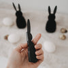 Schwarzer Papierhase in der Hand, weitere schwarze Papierhasen und Ostereier unscharf im Hintergrund.