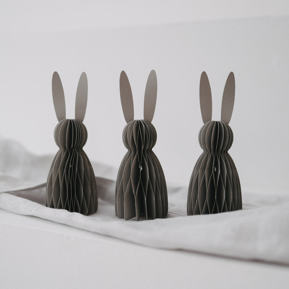 Drei graue Papierhasen im Honeycomb-Design als Osterdeko auf hellstem Stoff arrangiert.
