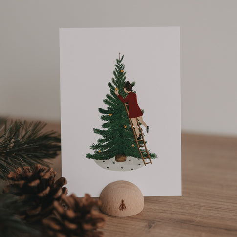Weihnachtskarte mit Illustration einer Frau, die einen Tannenbaum mit Sternen dekoriert