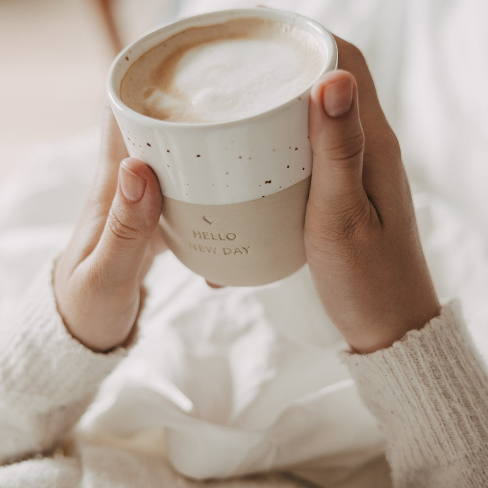 Beiger Keramikbecher „Hello New Day“ mit Herz und Milchschaum, in beiden Händen gehalten