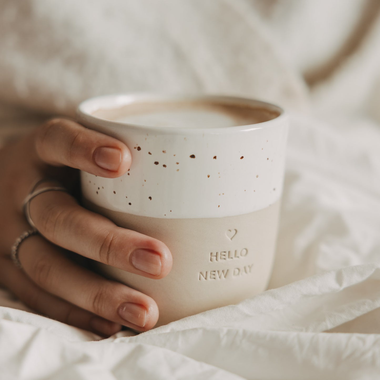 Beiger Keramikbecher „Hello New Day“ mit weiß gesprenkelter Glasur und Kaffee in der Hand