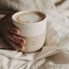 Keramikbecher „Hello New Day“ in Beige und Weiß mit Kaffee, gemütlich im Bett gehalten
