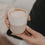 Nahaufnahme eines zweifarbigen Keramikbechers „Me Time“ mit Herz und Cappuccino, der in beiden Händen gehalten wird