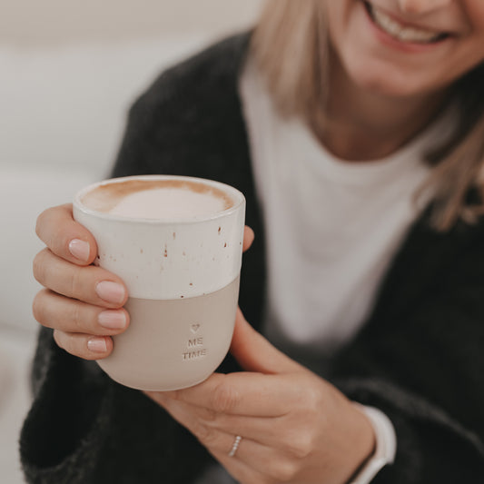 Lachende Frau hält einen zweifarbigen Keramikbecher „Me Time“ mit Herz und Cappuccino in den Händen