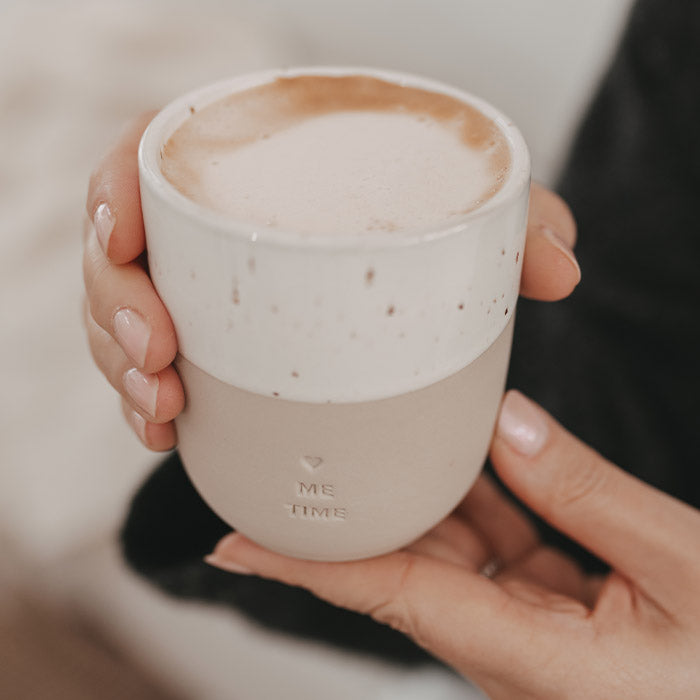 Nahaufnahme eines zweifarbigen Keramikbechers „Me Time“ mit Herz und Cappuccino, der in beiden Händen gehalten wird