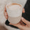Nahaufnahme eines zweifarbigen Keramikbechers „Me Time“ mit Herz und Cappuccino, der in beiden Händen gehalten wird