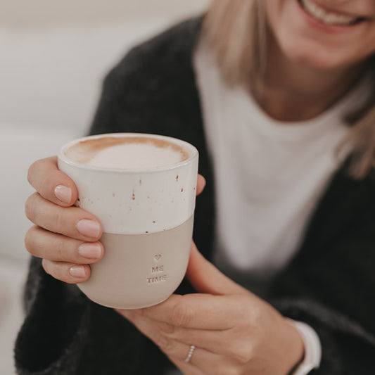 Lachende Frau hält einen zweifarbigen Keramikbecher „Me Time“ mit Herz und Cappuccino in den Händen
