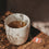 Beige-weißer Keramikbecher mit der Prägung „Glücklich“, gefüllt mit Tee und Teebeutel, im warmen Sonnenlicht auf einem Holztisch.