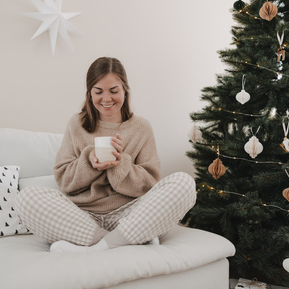 Lächelnde Frau sitzt gemütlich im Schneidersitz auf einem weißen Sofa und hält einen Keramikbecher von Eulenschnitt in den Händen. Neben ihr steht ein festlich geschmückter Weihnachtsbaum mit warmen Lichterketten und Papieranhängern.