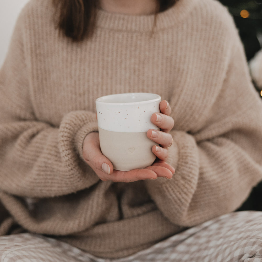 Frau in einem kuscheligen beigen Strickpullover hält einen zweifarbigen Keramikbecher von Eulenschnitt mit weiß gesprenkelter Glasur und kleiner Herzprägung in beiden Händen.