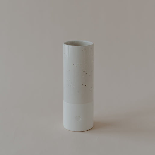 Zylindrische Vase aus Keramik mit gesprenkelter, glänzender Oberfläche im oberen Bereich und matter, weißer Oberfläche unten. Am unteren Teil ist ein kleines Herzrelief eingearbeitet. Die Vase steht auf hellem Untergrund vor neutralem Hintergrund.