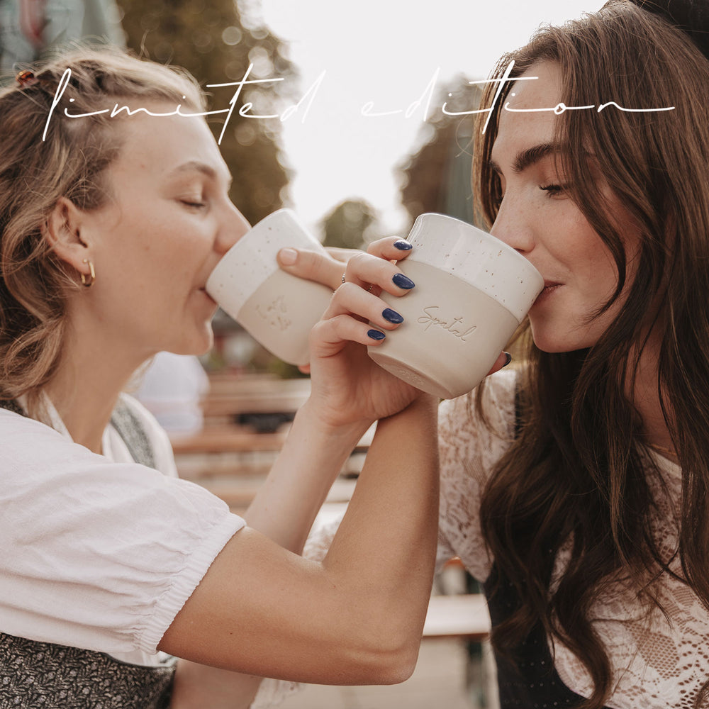 Zwei Frauen im Dirndl stoßen mit den limitierten Bechern „I mog di“ und „Spatzl“ beim Trinken an.