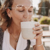 Frau im Dirndl trinkt aus dem Becher „I mog di“ und genießt den Moment in der Sonne.