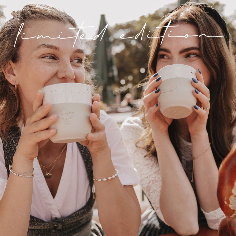 Zwei Frauen im Dirndl genießen die limitierten Becher „I mog di“ und „Spatzl“ im Biergarten.