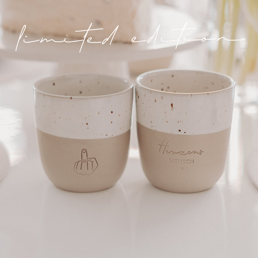 Zwei Limited Edition Keramikbecher in Beige und Weiß mit geprägten Motiven stehen nebeneinander auf einem Tisch.