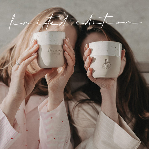 Zwei Frauen halten Herzensmensch- und Mittelfinger-Becher der Limited Edition vor sich, beige-weiß mit Keramikstruktur.