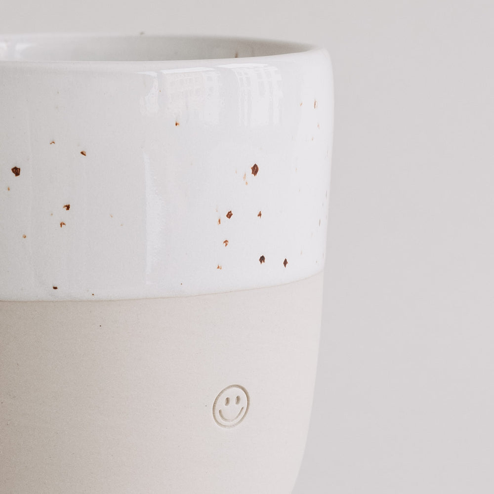 Keramikbecher in Naturtönen mit weißer Sprenkelglasur und kleinem Smiley-Relief, Nahaufnahme des oberen Randes.