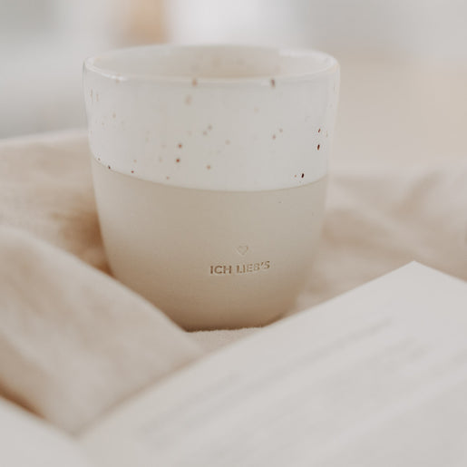 Beige-weiße „Ich lieb’s“-Tasse mit Sprenkelglasur auf Stoff, neben geöffnetem Buch.