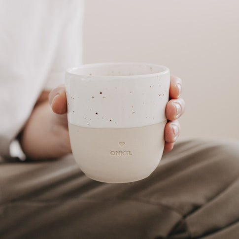 Person hält den Keramikbecher „Onkel“ mit gesprenkelter Glasur und Herzprägung in der Hand.