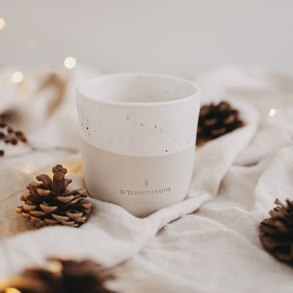 Becher „O Tannenbaum“ in weihnachtlicher Deko mit Zapfen und Lichterkette auf Stoff arrangiert.