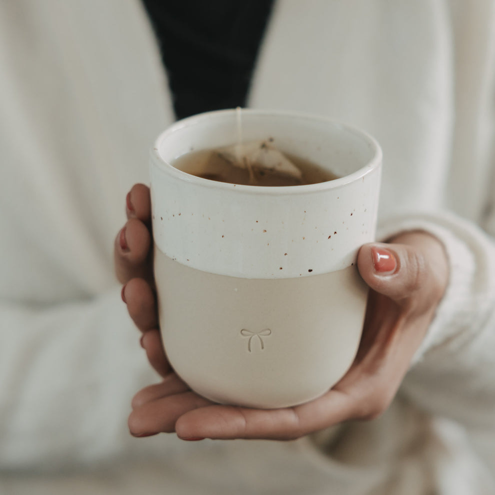 Keramikbecher mit Schleifenmotiv in Händen, gefüllt mit Tee, minimalistisches Design in warmem Licht