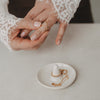 Weiße Keramik-Schmuckablage mit Ringhalter, Kette und Ringen neben Händen mit Goldring